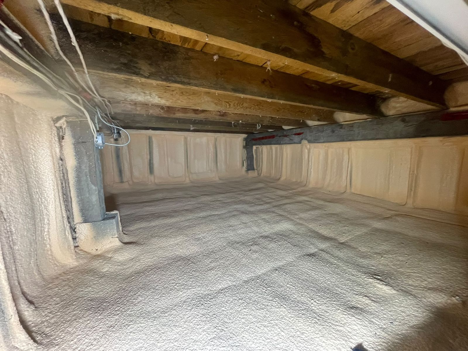 Crawl space with closed-cell spray foam fully encapsulating the perimeter walls and ground, floor joists and a wrapped support post visible above
