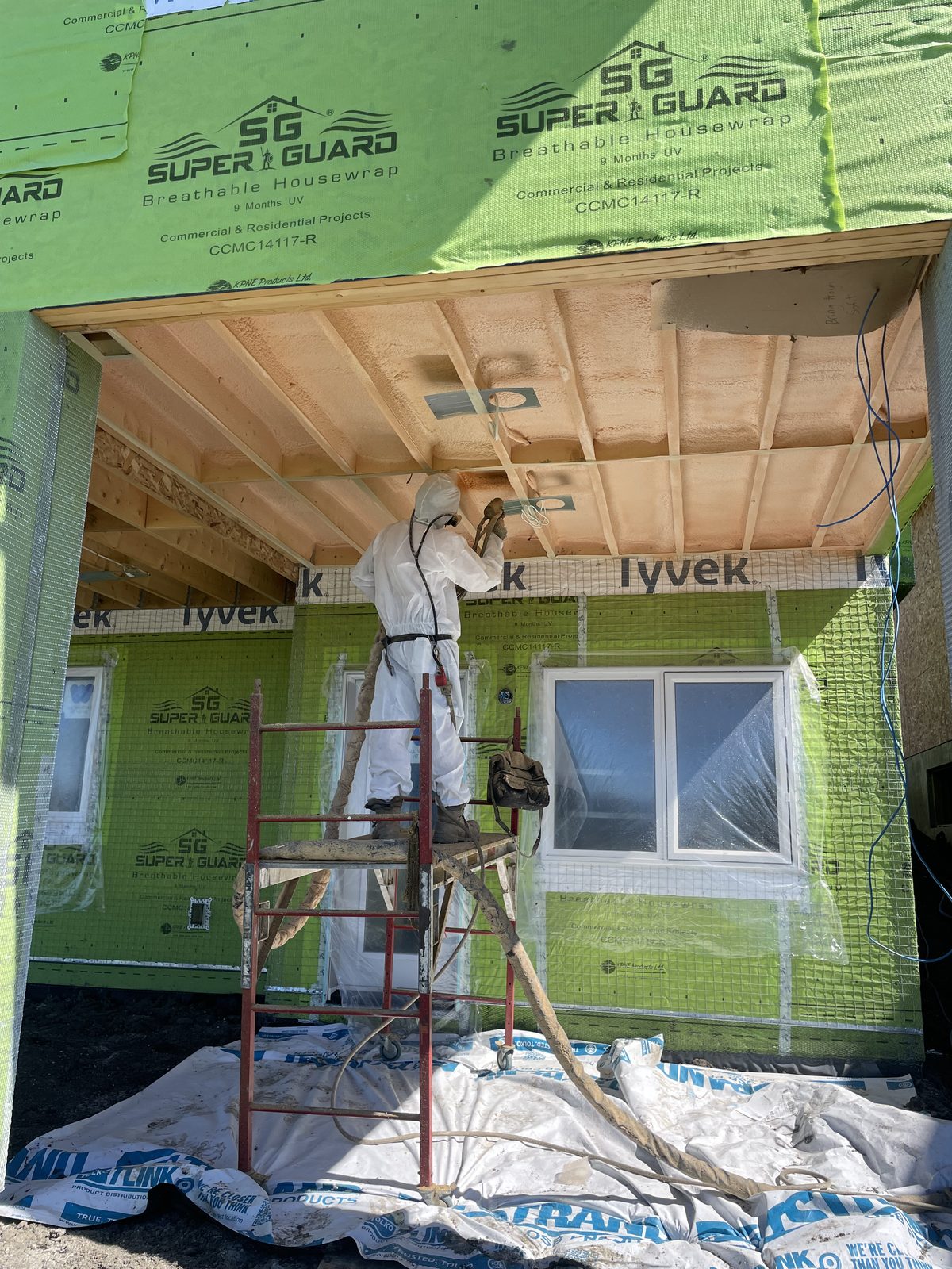 Installer on scaffolding spray foaming an exterior wall over housewrap, crew on site mid-application