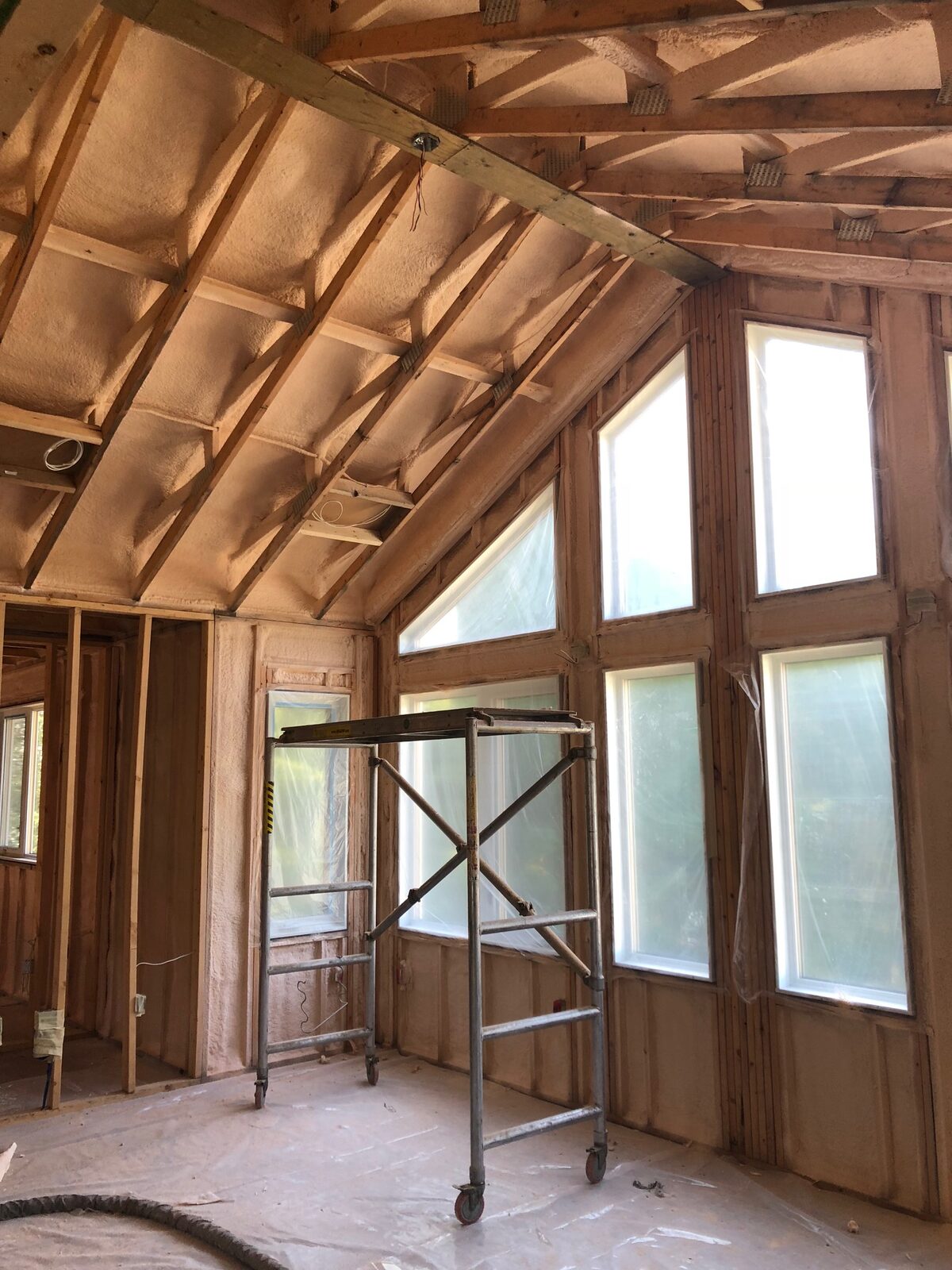 New-construction great room in St. Clements with closed-cell spray foam between the vaulted ceiling rafters, a tall window wall and a rolling scaffold on poly-covered subfloor