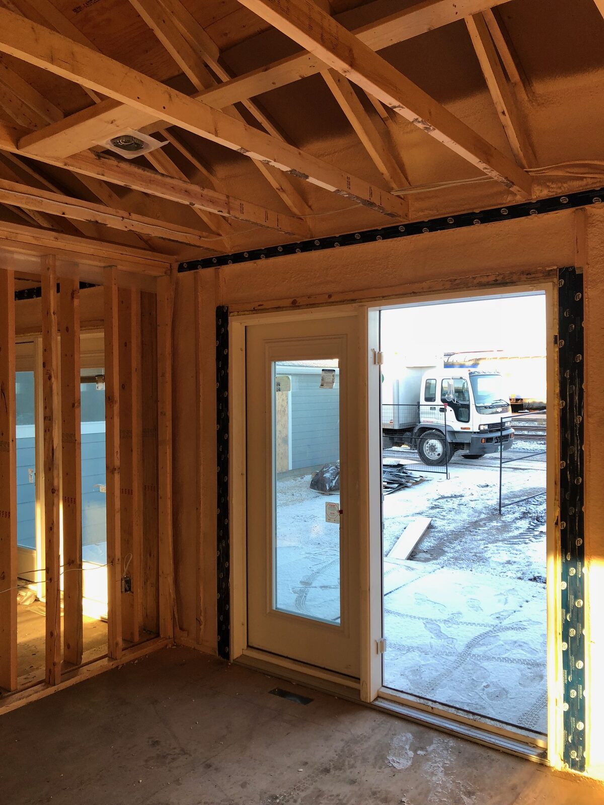 Double-stud wall in a Thompson housing unit with closed-cell foam between studs and black tape sealing the top plate and patio door opening, snow and a truck visible outside
