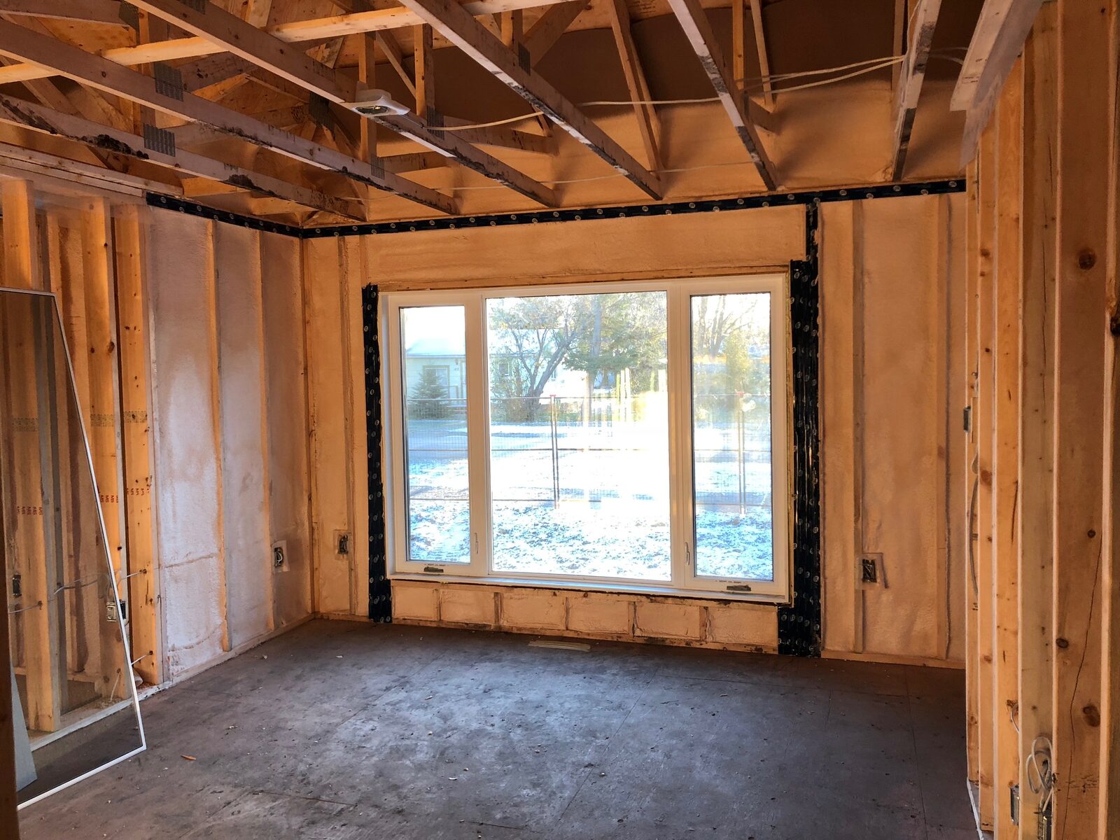 Living room wall with a large picture window, foamed stud bays and taped window jambs under open ceiling trusses