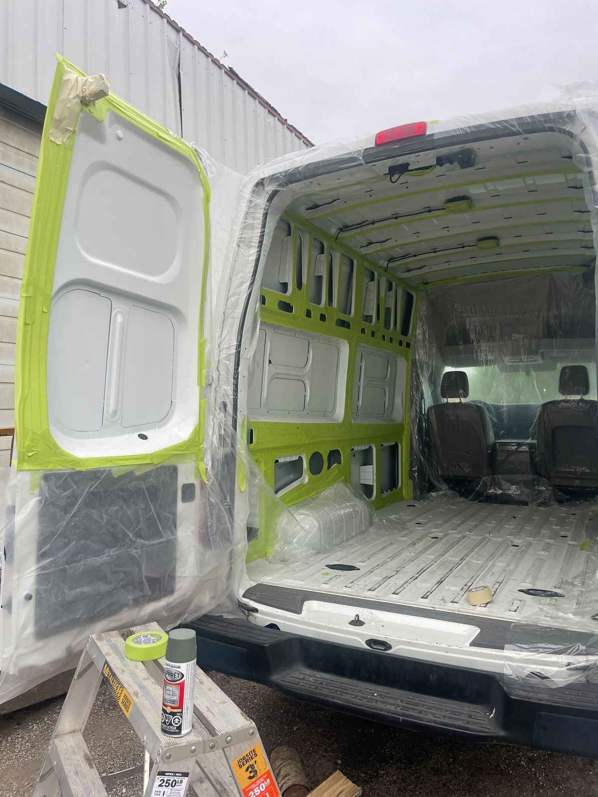 Cargo van interior masked off with tape and plastic sheeting, walls primed white, ready for spray foam insulation