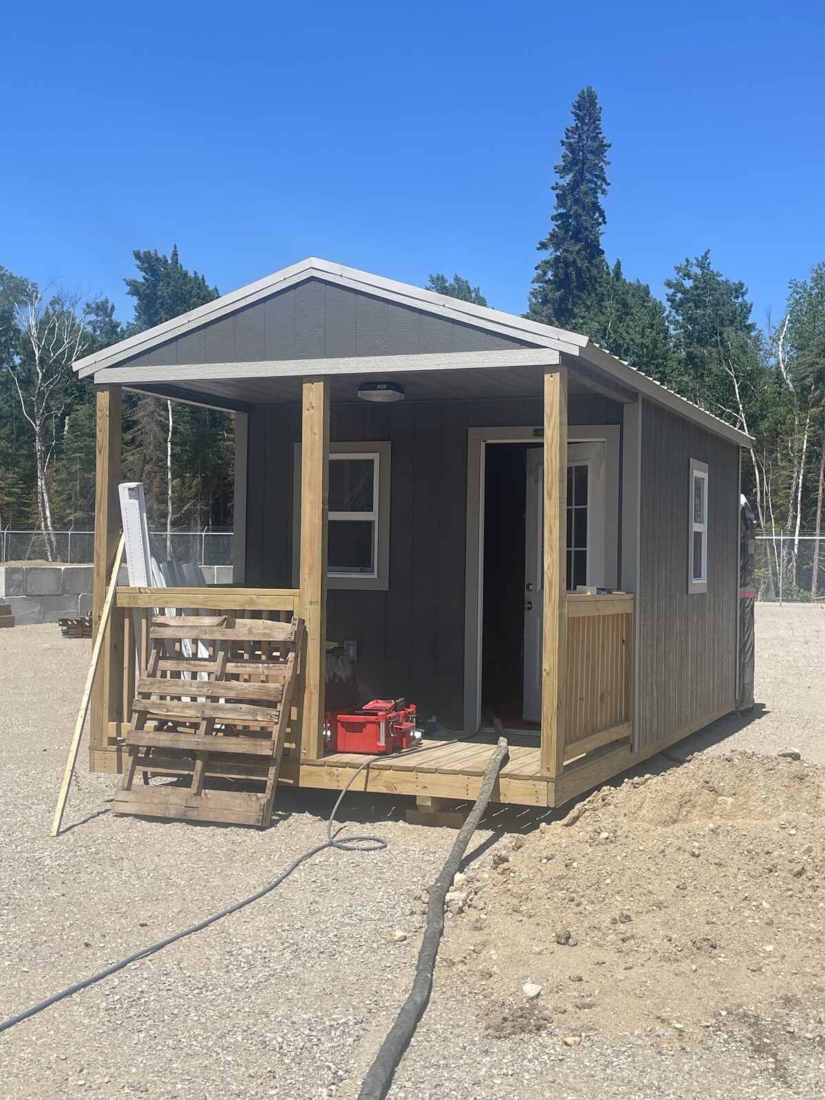 Small grey gable-roofed staff bunkie with a covered wooden porch on a gravel pad at the Victoria Beach garbage station, chain-link fence and spruce trees behind, spray hose running in the door